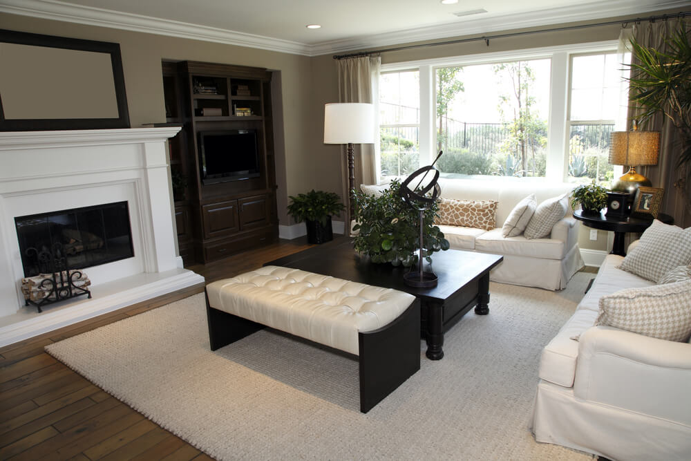 White and dark brown living room design. In addition to two all-white sofas, there is a low dark wood coffee table and dark wood and white leather bench. Furniture faces a large white fireplace. White and dark brown living room design. In addition to two all-white sofas, there is a low dark wood coffee table and dark wood and white leather bench. Furniture faces a large white fireplace.