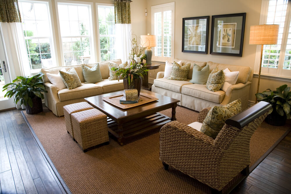 Contemporary living room design with two matching off-white sofas loaded with white and soft green pillows. One wicker armchair. Square coffee table supplemented with two small wicker ottomans. Contemporary living room design with two matching off-white sofas loaded with white and soft green pillows. One wicker armchair. Square coffee table supplemented with two small wicker ottomans.