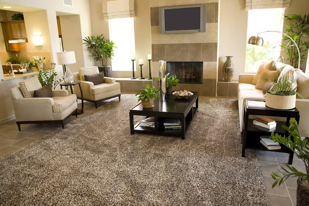 Contemporary living room design in an open concept home. Fireplace with mounted flat screen television is off to the side. Two armchairs face a sofa. Large rug creates texture on the tile floor. Contemporary living room design in an open concept home. Fireplace with mounted flat screen television is off to the side. Two armchairs face a sofa. Large rug creates texture on the tile floor.