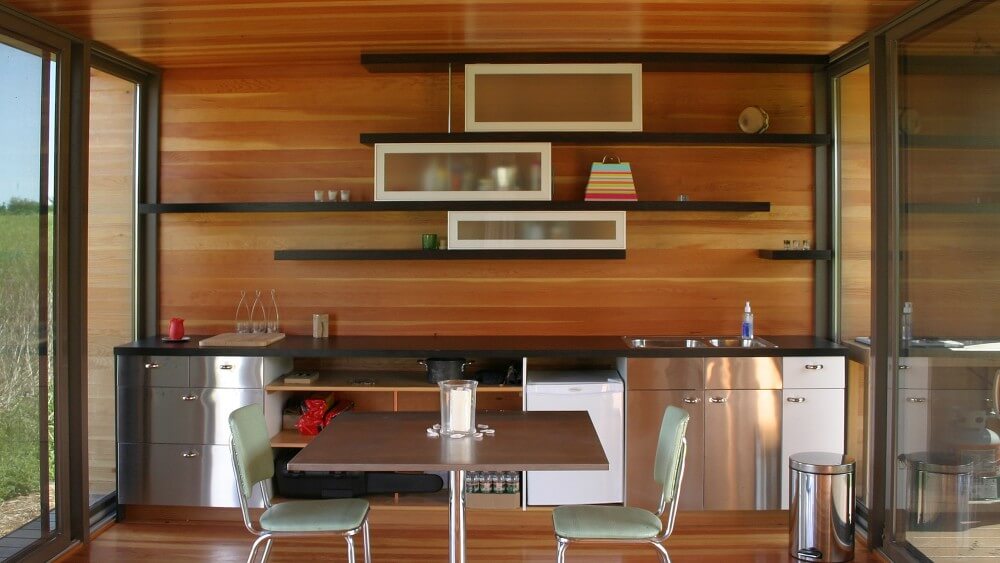 Kitchen side of the home features unique built-in asymmetrical black shelving with white glass box cupboards over aluminum cupboards and black countertop. Small dining table rests at center with blue chairs. Kitchen side of the home features unique built-in asymmetrical black shelving with white glass box cupboards over aluminum cupboards and black countertop. Small dining table rests at center with blue chairs.