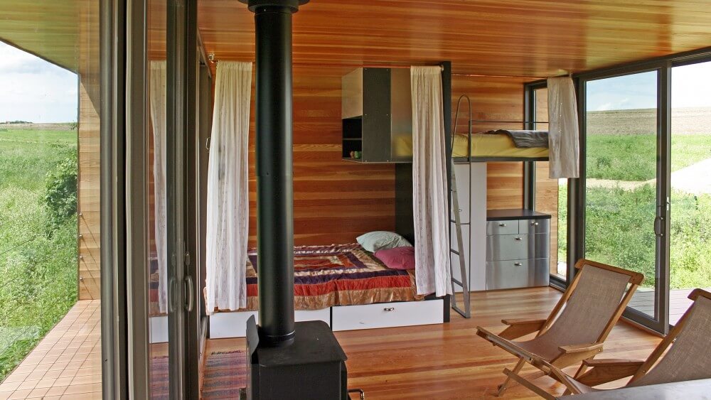 Sleeping quarters features small bunk bed over brushed aluminum dresser next to full bed with built in storage below and above. Pot bellied stove and chairs share the space. Sleeping quarters features small bunk bed over brushed aluminum dresser next to full bed with built in storage below and above. Pot bellied stove and chairs share the space.
