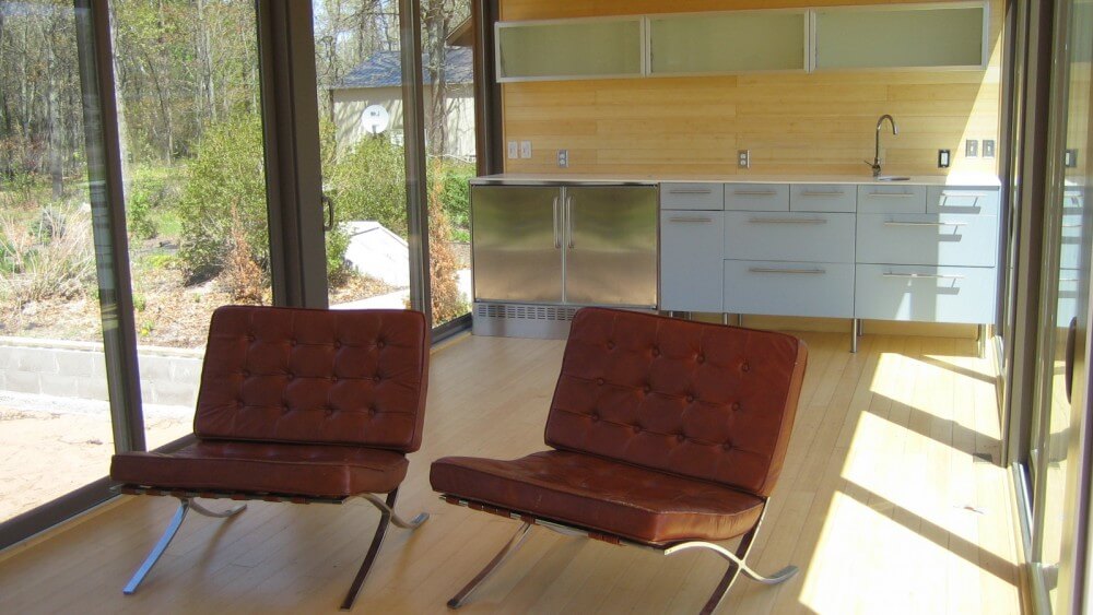 Opposite view reveals kitchen area with white and metal cabinetry, glass door shelves mounted on wall, and twin leather living room chairs in foreground. Opposite view reveals kitchen area with white and metal cabinetry, glass door shelves mounted on wall, and twin leather living room chairs in foreground.