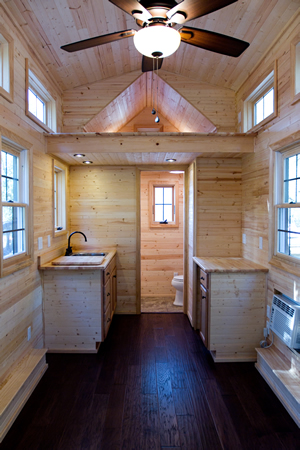 Interior of tiny home maximizes space, with small bathroom next to kitchenette. Light natural wood over dark hardwood flooring throughout. Interior of tiny home maximizes space, with small bathroom next to kitchenette. Light natural wood over dark hardwood flooring throughout.