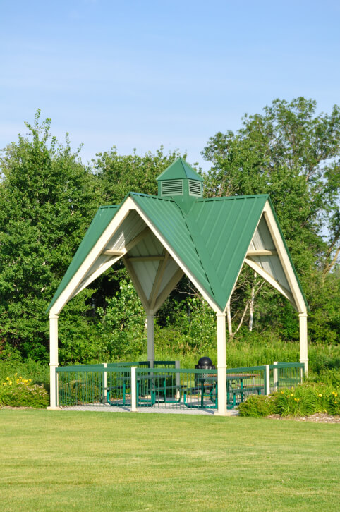 A tall gable roof in a bright green makes this public gazebo an easy structure to spot from a long ways away. The gazebo is furnished with a picnic table, chairs, and a trash can. A tall gable roof in a bright green makes this public gazebo an easy structure to spot from a long ways away. The gazebo is furnished with a picnic table, chairs, and a trash can.