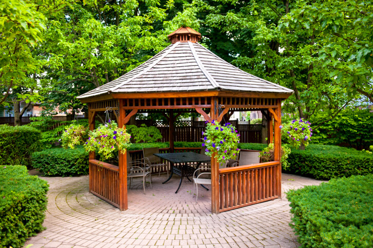 This cedar gazebo is home to a small patio dining set and is placed at the center of a garden on a brick patio surrounded by hedges and trees. This cedar gazebo is home to a small patio dining set and is placed at the center of a garden on a brick patio surrounded by hedges and trees.