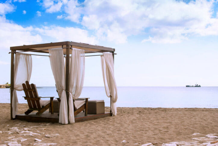 A square pergola-style gazebo fitted with sheer curtains and adirondack chairs. The gazebo looks out on the water and embraces a beachy aesthetic. A square pergola-style gazebo fitted with sheer curtains and adirondack chairs. The gazebo looks out on the water and embraces a beachy aesthetic.