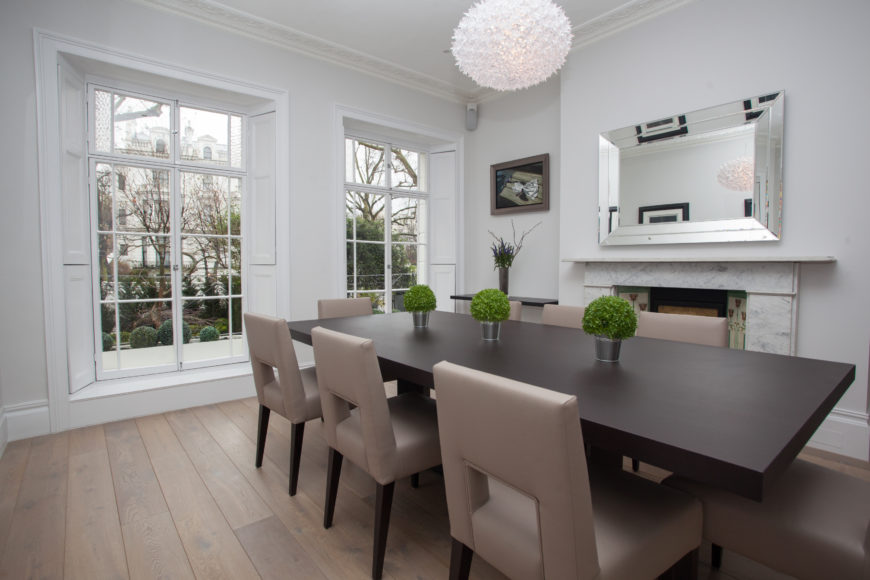 Formal dining room designed with a fancy pendant light and rectangular mirror mounted above the fireplace. It has a wooden dining table and taupe chairs over wood plank flooring.