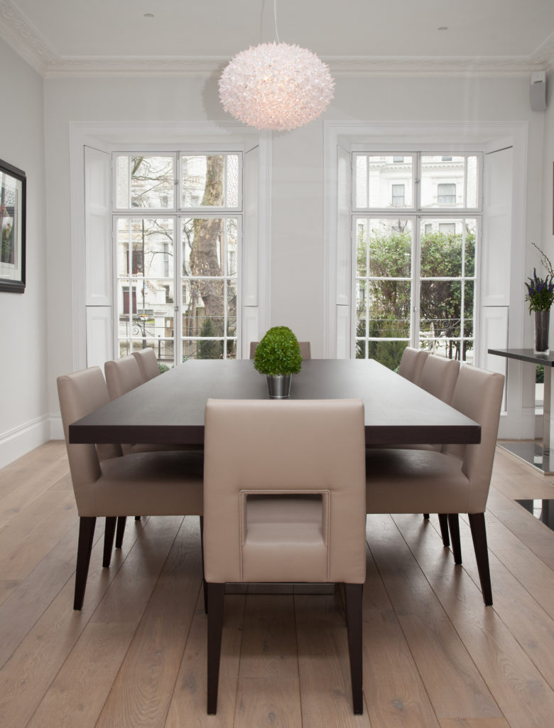 A classic dining room highlighted by a charming crystal chandelier along with light hardwood flooring. It is framed with light gray walls and white glass windows.