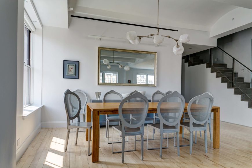 A dining area with a rectangular wood table surrounded by gray round back dining chairs. It is decorated with a wall mirror mounted above the buffet table and a modern chandelier.