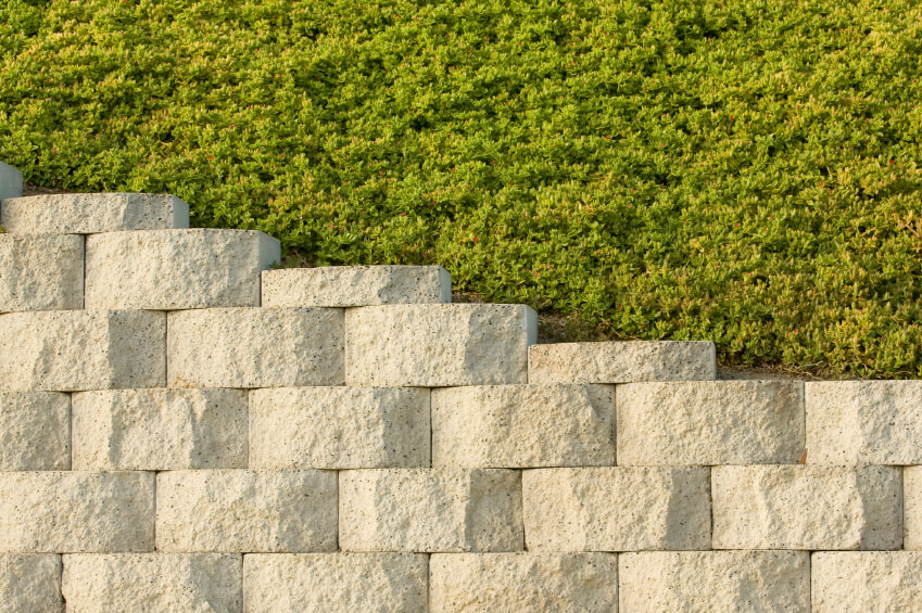 A retaining wall of light stacked stone that is uniform in color and size. The ground above the stones is covered in a thick groundcover. A retaining wall of light stacked stone that is uniform in color and size. The ground above the stones is covered in a thick groundcover.