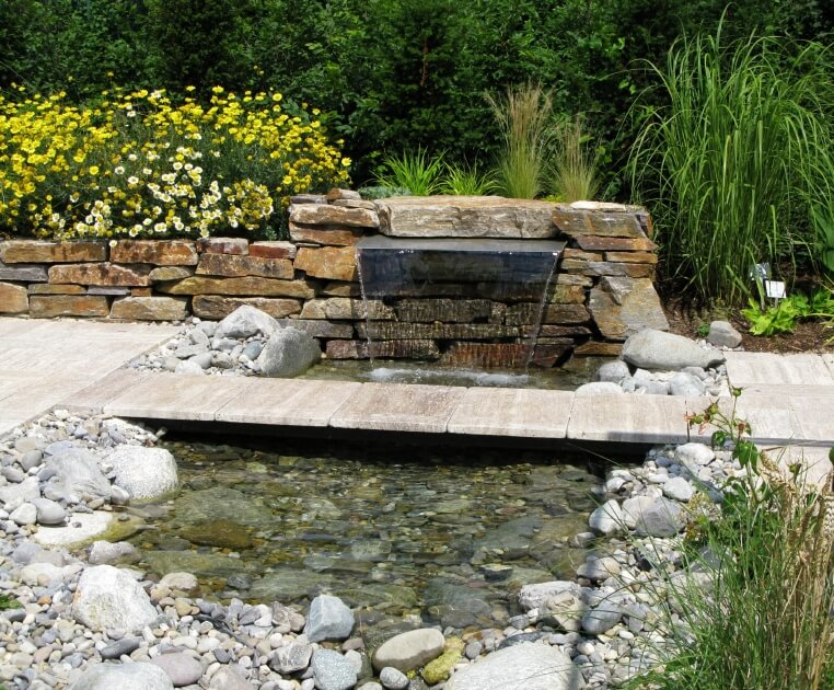 A shallow pond fed by a cascading fountain tucked into the stone edging. A narrow stone footbridge connects the two sides of the pathway. A shallow pond fed by a cascading fountain tucked into the stone edging. A narrow stone footbridge connects the two sides of the pathway.