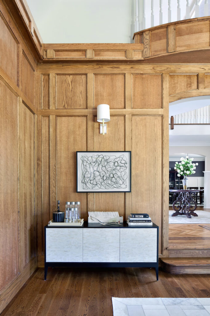 The den entrance is off of the foyer, and immediately to the right of the entrance is a small buffet area with a stunning piece of contemporary art. Visible through the archway is the ornate dark wood table with a vase of white flowers that brightens the foyer.