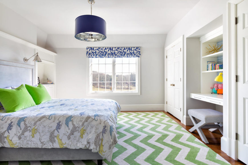 A child's bedroom in white and green, featuring a fun dinosaur bedspread. The area rug is a chevon pattern in a light, springy green. The secondary accent color is a bold deep blue, in the light fixture and on the window coverings.