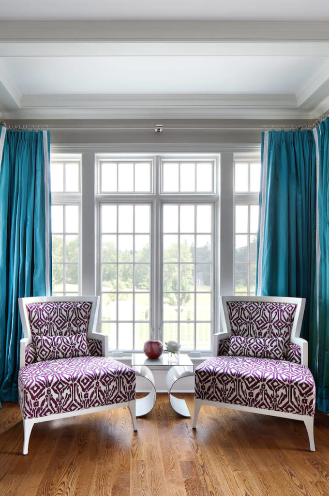  A closer look at the sitting room in white and purple in front of the large windows. Between the two chairs is a modern side table with a few small decorative pieces. The bold turquoise drapes behind the chairs is a bold statement in color.
