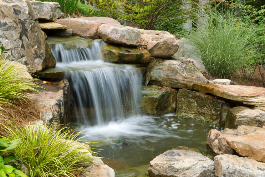 This water feature focuses on the large waterfall tumbling into a small, shallow pool at the bottom. The entire structure is encased by large stones and surrounded by grasses. This water feature focuses on the large waterfall tumbling into a small, shallow pool at the bottom. The entire structure is encased by large stones and surrounded by grasses.
