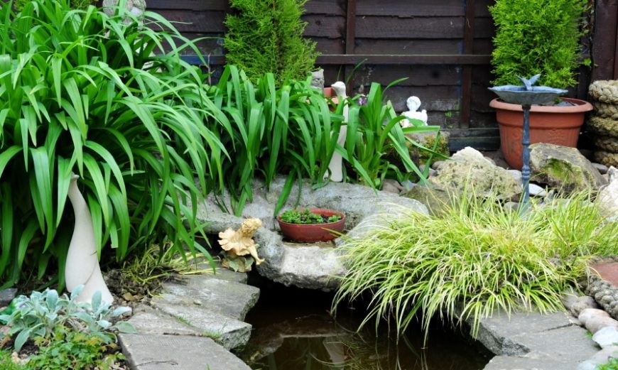 A well-like garden pond with a small fountain that pours into the deeper well of water. Thick grasses drape over the stone edges. The garden is filled with stone statues and other accents. A well-like garden pond with a small fountain that pours into the deeper well of water. Thick grasses drape over the stone edges. The garden is filled with stone statues and other accents.