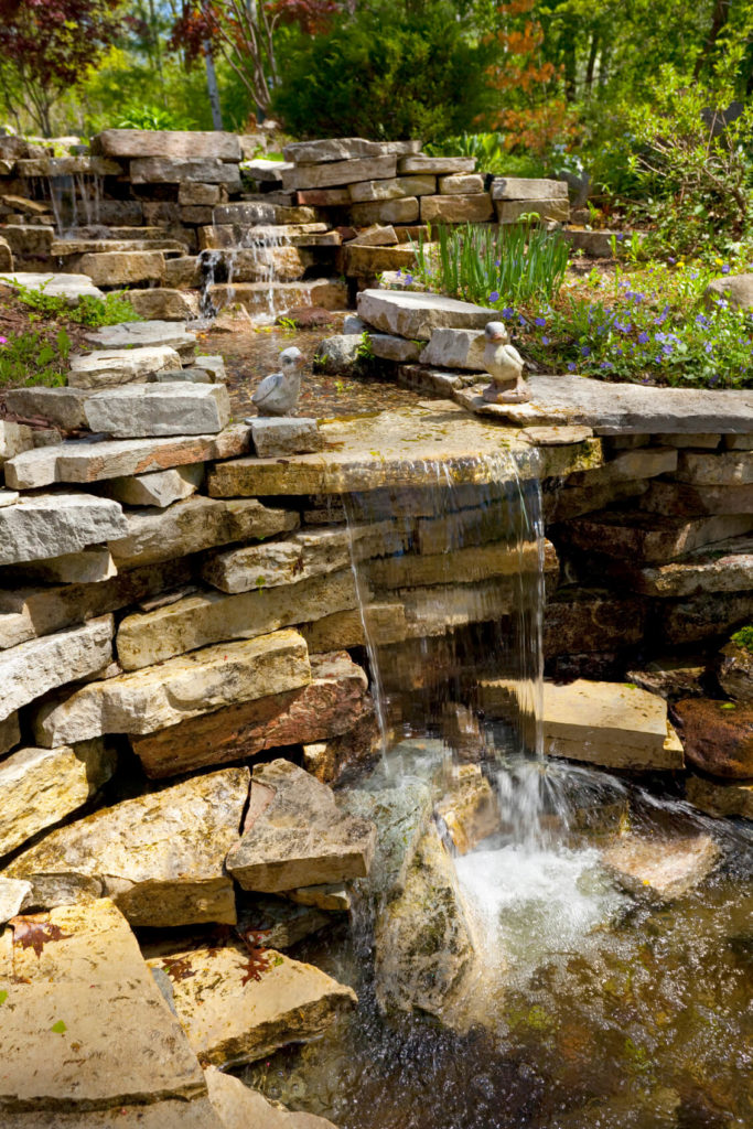 An awe-inspiring stone waterfall pouring into a shallow basin at the bottom. Small figures of ducks sit on either side of the largest cascade. An awe-inspiring stone waterfall pouring into a shallow basin at the bottom. Small figures of ducks sit on either side of the largest cascade.
