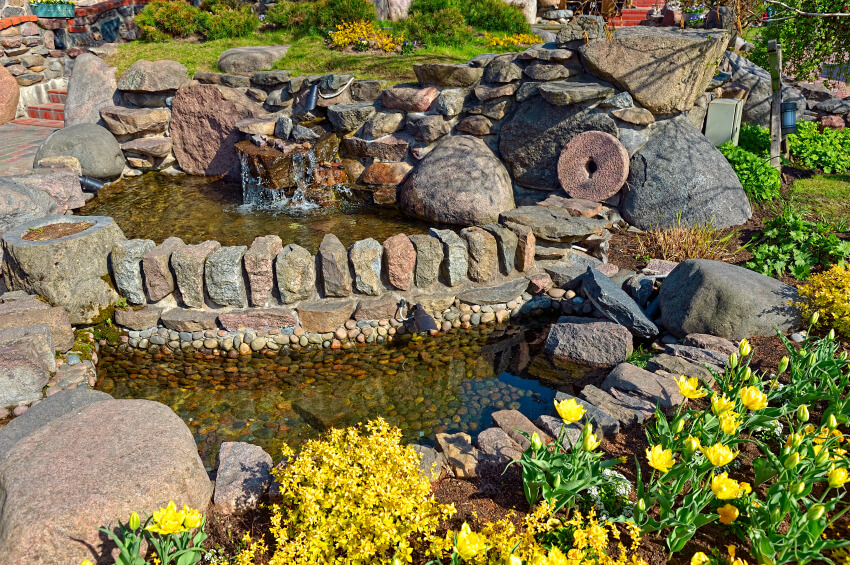 A unique pond with two tiers. The top tier has a small rock waterfall. Fields of tulips and other plants surround the rock enclosure. A unique pond with two tiers. The top tier has a small rock waterfall. Fields of tulips and other plants surround the rock enclosure.