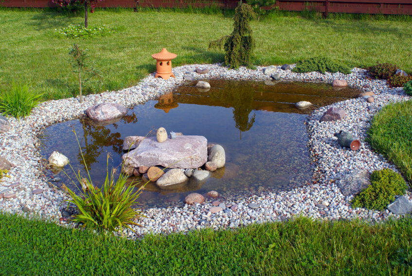 A shallow garden pool with a few ornamental rocks and grasses along the pebble edging. A small terra cotta Japanese lantern sits on one edge. A shallow garden pool with a few ornamental rocks and grasses along the pebble edging. A small terra cotta Japanese lantern sits on one edge.