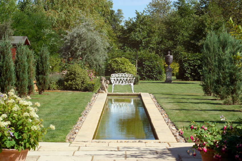 A more cosmopolitan pool stretching out into the yard from the patio and ending in a white bench and angle statue. Evergreens and other large trees and bushes line either side of the yard. A more cosmopolitan pool stretching out into the yard from the patio and ending in a white bench and angle statue. Evergreens and other large trees and bushes line either side of the yard.