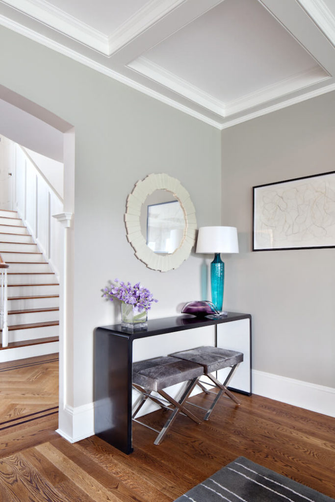 A small desk area with velvet seating and glass vase decorations sits on the other side of the doorframe from the shelving unit. Above the desk is a circular mirror in ivory. The table lamp is in a beautiful bold turquoise.