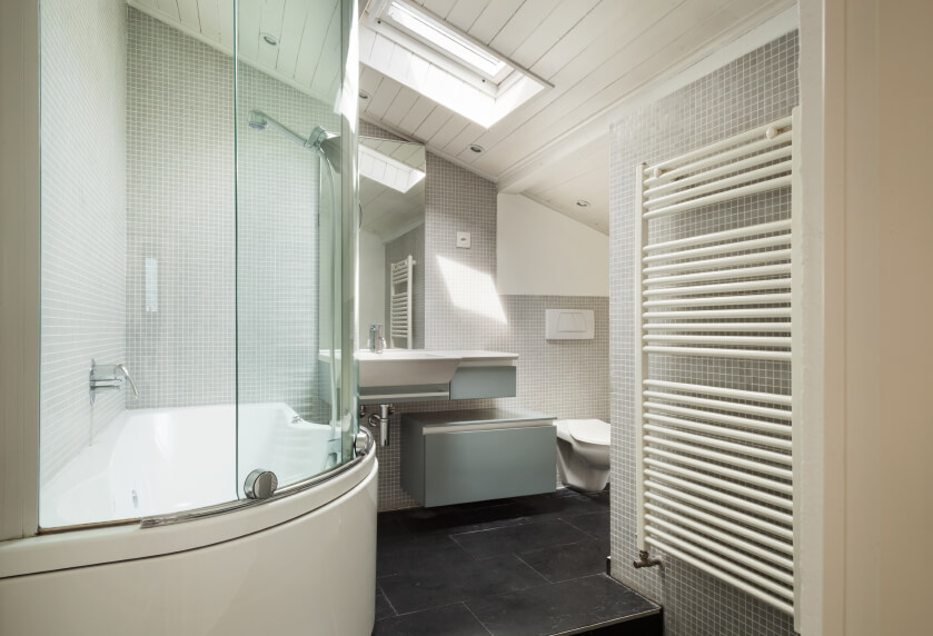 A modern industrial style bathroom features a rounded bathing area, heated towel rack, and petite gray tiles complementing the charcoal stone tiling of the floor. A skylight beams down from the angled planked ceiling, and a modern tankless toilet and sink round out this appealing bathroom. A modern industrial style bathroom features a rounded bathing area, heated towel rack, and petite gray tiles complementing the charcoal stone tiling of the floor. A skylight beams down from the angled planked ceiling, and a modern tankless toilet and sink round out this appealing bathroom.