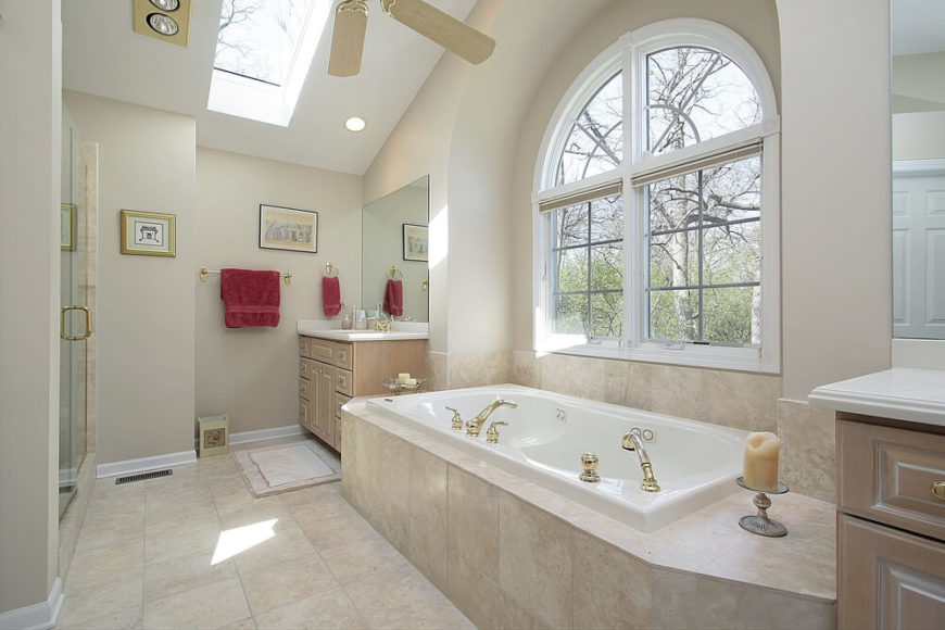 This neutral palette bathroom features stone tile-work, two vanities, and a lovely arched window allowing views of the outside world. A ceiling skylight allows for additional light flow in this already bright space, while maroon accent towels contribute vibrant color to an otherwise muted space. This neutral palette bathroom features stone tile-work, two vanities, and a lovely arched window allowing views of the outside world. A ceiling skylight allows for additional light flow in this already bright space, while maroon accent towels contribute vibrant color to an otherwise muted space.