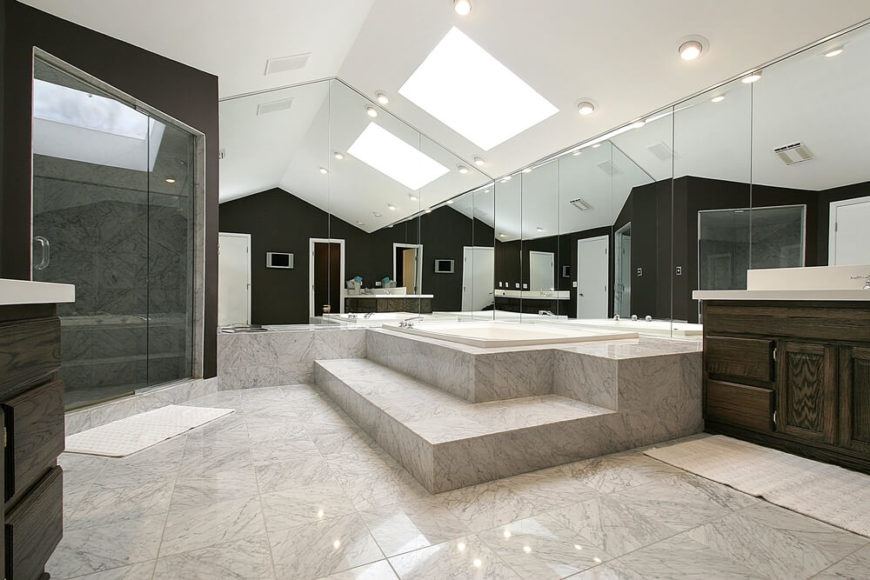 This stunning bathroom features glossy squares of stone tile, soaring ceilings, and wall length mirrors contributing to the extraordinary illusion of increased space. A skylight hangs above, reflecting off mirrors and allowing light to bounce around the room. Dark walls and cabinetry offer stark contrast to the muted tones of the rest of this impressive space. This stunning bathroom features glossy squares of stone tile, soaring ceilings, and wall length mirrors contributing to the extraordinary illusion of increased space. A skylight hangs above, reflecting off mirrors and allowing light to bounce around the room. Dark walls and cabinetry offer stark contrast to the muted tones of the rest of this impressive space.