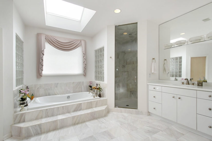 This light space features a tinted glass enclosed shower, a beautiful skylight, and sizable mirror allowing reflection of light and illusion of space. The simple white cabinetry complements the light stone tiles seen throughout the room, while a step leading into the relaxing spa bathtub offers convenience. This light space features a tinted glass enclosed shower, a beautiful skylight, and sizable mirror allowing reflection of light and illusion of space. The simple white cabinetry complements the light stone tiles seen throughout the room, while a step leading into the relaxing spa bathtub offers convenience.