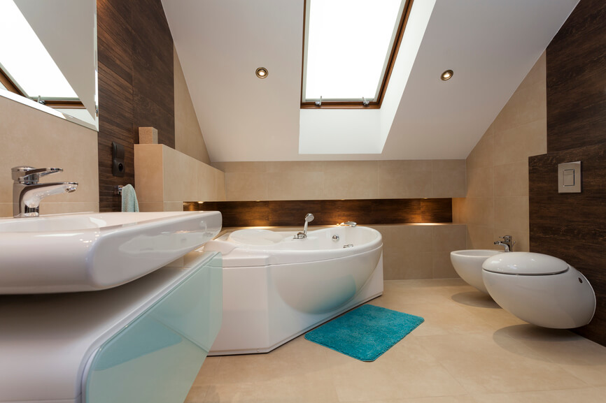 This wonderfully modern bathroom relies on an attractive skylight and recessed lighting to brighten the space. The gentle curves of the bathtub, sink, and bidet are eye catching and attractive. A lone turquoise bathmat offers a welcome splash of bold color against the neutral base of the cream tile flooring. This wonderfully modern bathroom relies on an attractive skylight and recessed lighting to brighten the space. The gentle curves of the bathtub, sink, and bidet are eye catching and attractive. A lone turquoise bathmat offers a welcome splash of bold color against the neutral base of the cream tile flooring.