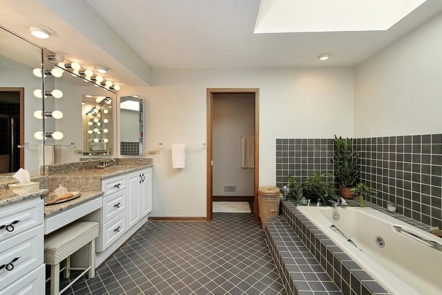 Black tiles with white grouting is attractive and eye catching in this wonderful bathroom space. The double vanity features a seating area, and soft lightbulbs offer the vintage ambiance of an old Hollywood dressing room. A skylight looms above the narrow tub, with decorative greenery strategically placed to provide beauty and additional texture. Black tiles with white grouting is attractive and eye catching in this wonderful bathroom space. The double vanity features a seating area, and soft lightbulbs offer the vintage ambiance of an old Hollywood dressing room. A skylight looms above the narrow tub, with decorative greenery strategically placed to provide beauty and additional texture.