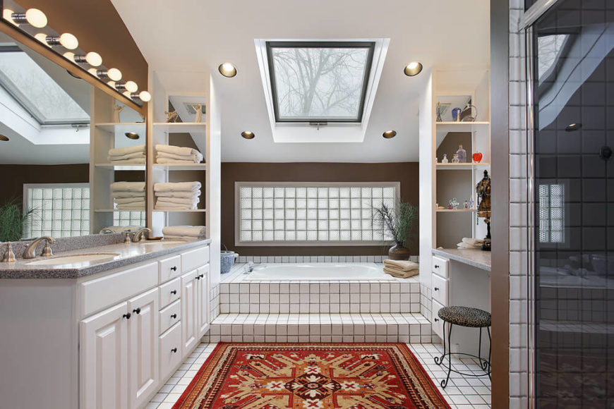This bathroom is rich in patterns,contrast, and textures that create a dynamic and interesting space. Simple square tiles with dark grouting are mimicked in the frosted glass panel above the bathtub area. A skylight shines down, aided by recessed lighting and a simple lighting fixture above the mirror. A double vanity and shelving offer plenty of storage, while a bright rust area rug leads the way to the tiled steps of the encased bathtub. This bathroom is rich in patterns,contrast, and textures that create a dynamic and interesting space. Simple square tiles with dark grouting are mimicked in the frosted glass panel above the bathtub area. A skylight shines down, aided by recessed lighting and a simple lighting fixture above the mirror. A double vanity and shelving offer plenty of storage, while a bright rust area rug leads the way to the tiled steps of the encased bathtub.