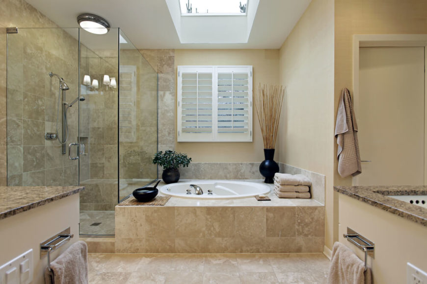 Simple country shutters filter in natural light, while a skylight allows sunshine to flood in. Additional lighting fixtures can be seen in the reflection of the glass enclosed shower, while cream vanities topped with dark marble offer a bit of contrast. Simple country shutters filter in natural light, while a skylight allows sunshine to flood in. Additional lighting fixtures can be seen in the reflection of the glass enclosed shower, while cream vanities topped with dark marble offer a bit of contrast.