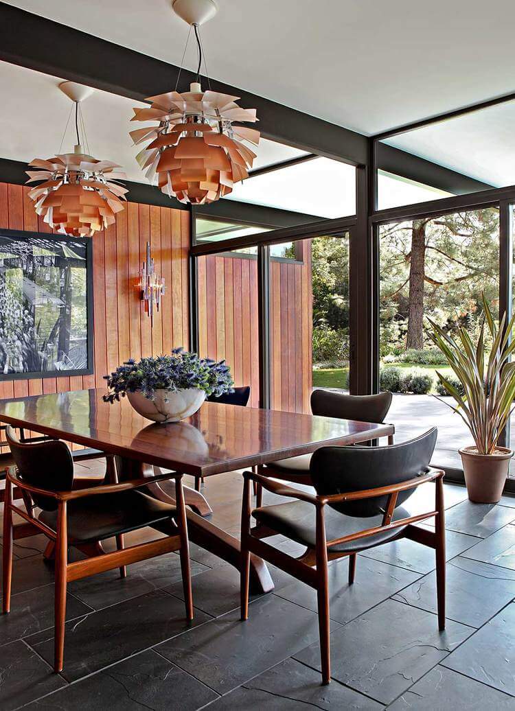Charming dining space with rustic wooden walls, rectangular wooden table, rustic wooden chairs, floor to ceiling windows, a stark contrast from its black tiles, and two interesting pendant lights.