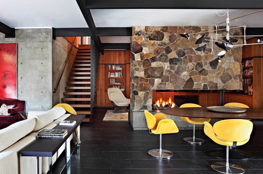 Cozy dining space with a fireplace, surprising pops of yellow colors from the unique chairs, round wooden table, and black floor tiles.