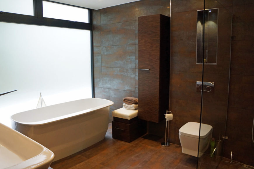 Contemporary primary bathroom accented by copper tones. It includes a freestanding tub near the frosted glass window and a toilet and shower area divided by tempered glass.