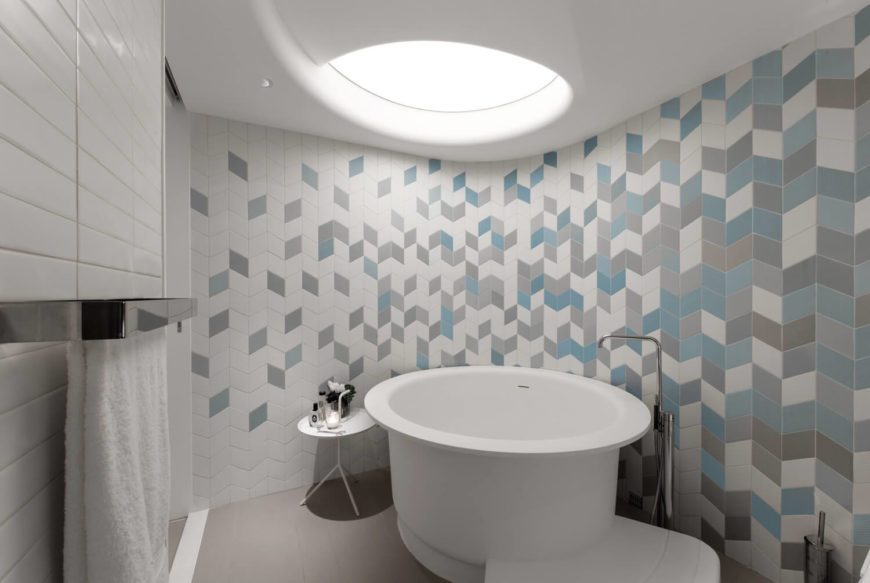An eye-catching chevron shower wall tile highlights this primary bathroom. It features a freestanding bathtub underneath a round skylight bringing natural light in.