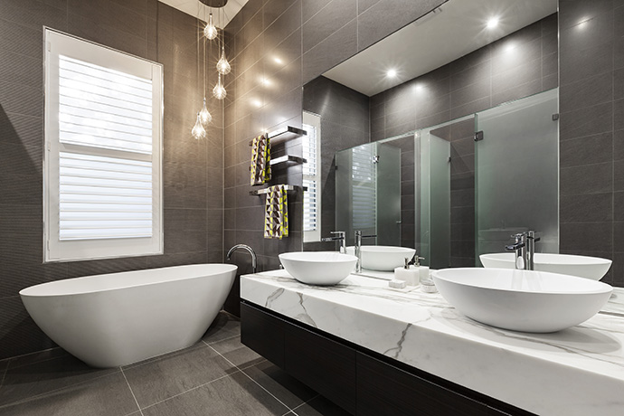 Cascading pendant lights illuminated the pedestal tub placed underneath a white framed window. This primary bathroom also features dual vessel sinks that sit on a white marble counter.