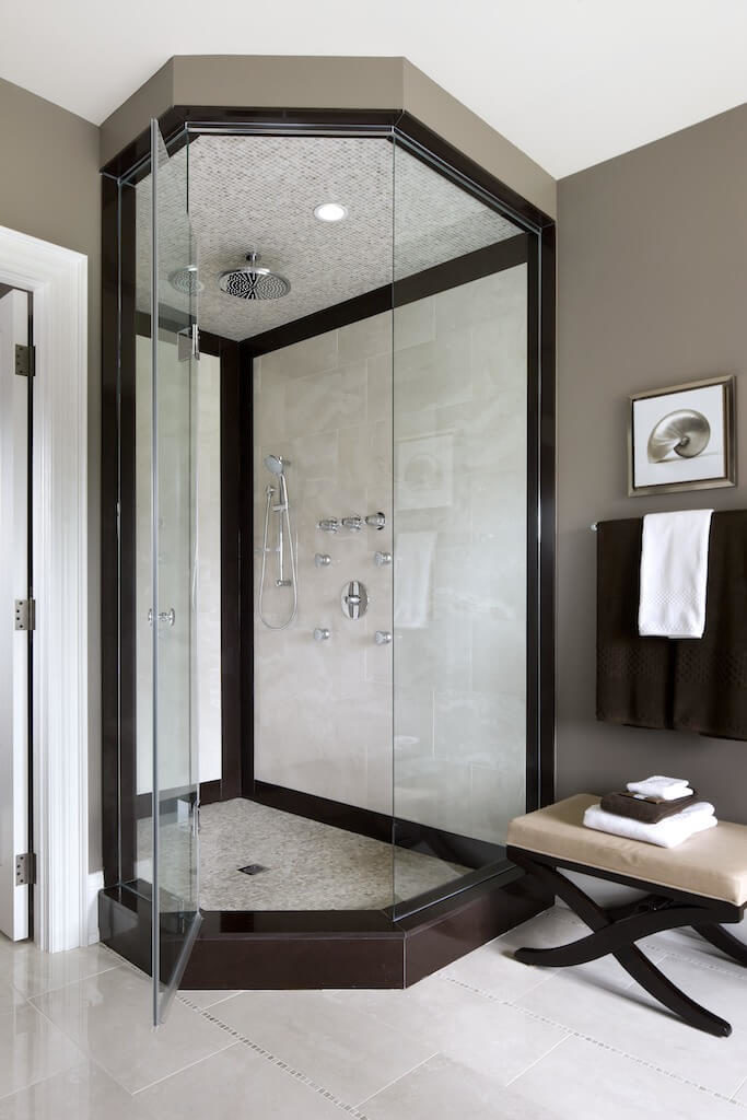 The same bathroom has an glass enclosed shower with an uncommon shape to it. The corner shower is encased with a dark wooden perimeter. The same bathroom has an glass enclosed shower with an uncommon shape to it. The corner shower is encased with a dark wooden perimeter.