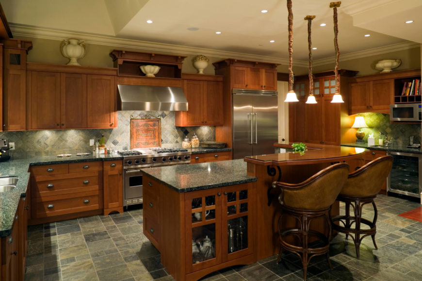 In this dark hued, utterly bespoke kitchen, sharp contrast is achieved via juxtaposing between dark floor tiling and matching-toned granite countertops against rich wood cabinetry. The island features glass door cabinetry and a two-tier design, with an upper level in rich wood for dining purposes. In this dark hued, utterly bespoke kitchen, sharp contrast is achieved via juxtaposing between dark floor tiling and matching-toned granite countertops against rich wood cabinetry. The island features glass door cabinetry and a two-tier design, with an upper level in rich wood for dining purposes.