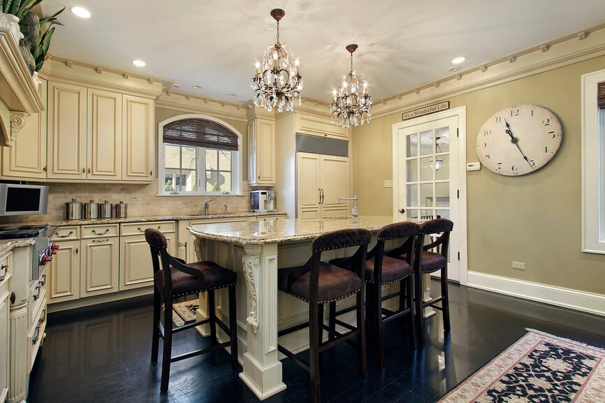 In a luxuriously detailed kitchen, high contrast informs the overall style. Dark hardwood flooring matches light beige cabinetry, while the granite topped island features ornate carved detail and abundant room for in-kitchen dining, courtesy of a set of leather upholstered bar stools. In a luxuriously detailed kitchen, high contrast informs the overall style. Dark hardwood flooring matches light beige cabinetry, while the granite topped island features ornate carved detail and abundant room for in-kitchen dining, courtesy of a set of leather upholstered bar stools.