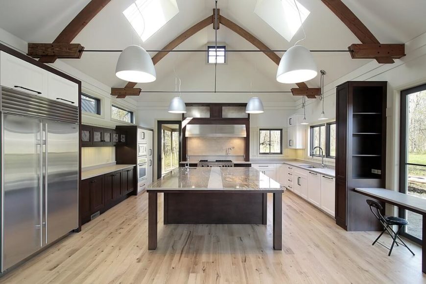 This bright kitchen stands below a massive vaulted ceiling with exposed natural wood beams, centered on a massive island with a full size dining table extension. The glass surface shows off the rich wood construction, which contrasts with the lighter hardwood flooring and white cabinetry throughout the kitchen. This bright kitchen stands below a massive vaulted ceiling with exposed natural wood beams, centered on a massive island with a full size dining table extension. The glass surface shows off the rich wood construction, which contrasts with the lighter hardwood flooring and white cabinetry throughout the kitchen.