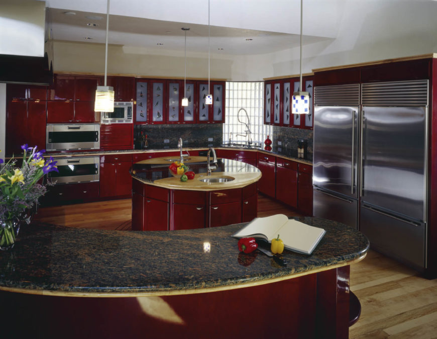 In a modern kitchen flush with bold red cabinetry, an innovative circular island holds the center. Wrapped in the same cherry red tone as the rest of the kitchen, the island sports a unique countertop, pairing granite with light wood. A set of dual sinks and wraparound cabinetry provide a surprising amount of utility in a small package. In a modern kitchen flush with bold red cabinetry, an innovative circular island holds the center. Wrapped in the same cherry red tone as the rest of the kitchen, the island sports a unique countertop, pairing granite with light wood. A set of dual sinks and wraparound cabinetry provide a surprising amount of utility in a small package.