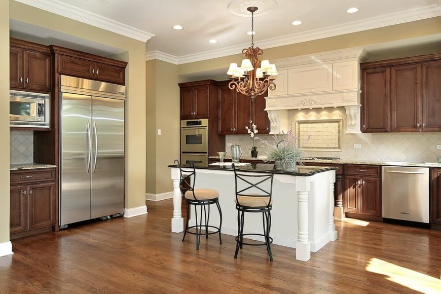 In an open but traditionally appointed kitchen wrapped in natural wood cabinetry, the elegant white island at center draws attention while subtly reflecting the white tile backsplash. In contrast with the hardwood flooring and beige granite countertops, a black countertop on the island truly punctuates the scene. In an open but traditionally appointed kitchen wrapped in natural wood cabinetry, the elegant white island at center draws attention while subtly reflecting the white tile backsplash. In contrast with the hardwood flooring and beige granite countertops, a black countertop on the island truly punctuates the scene.