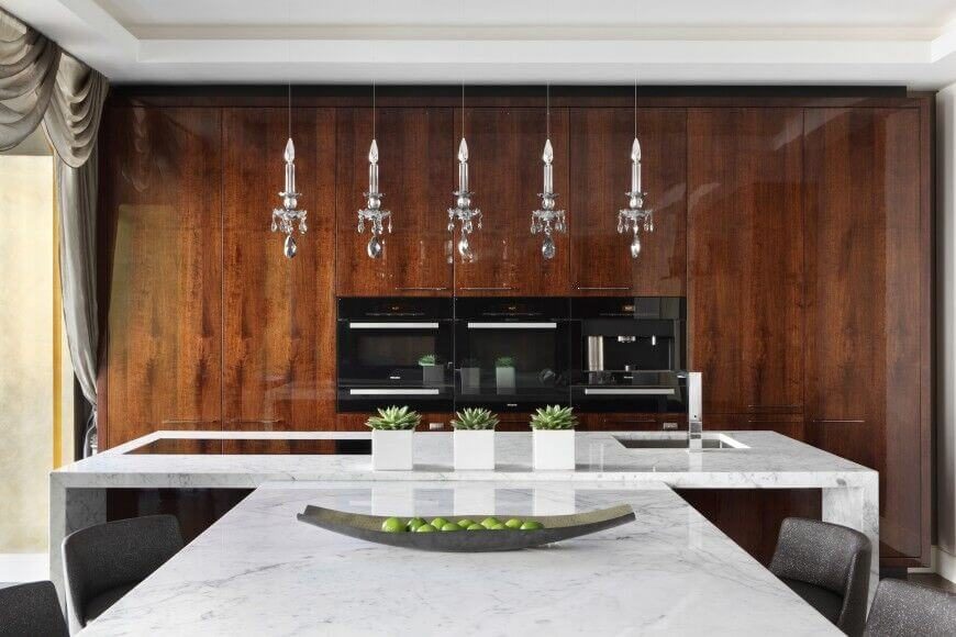 Here, slim appliances are nestled into a glossy wall of rich wood cabinets. A dining table in white marble extends directly from the island, and five crystal single candle lights hang above the island. Here, slim appliances are nestled into a glossy wall of rich wood cabinets. A dining table in white marble extends directly from the island, and five crystal single candle lights hang above the island.