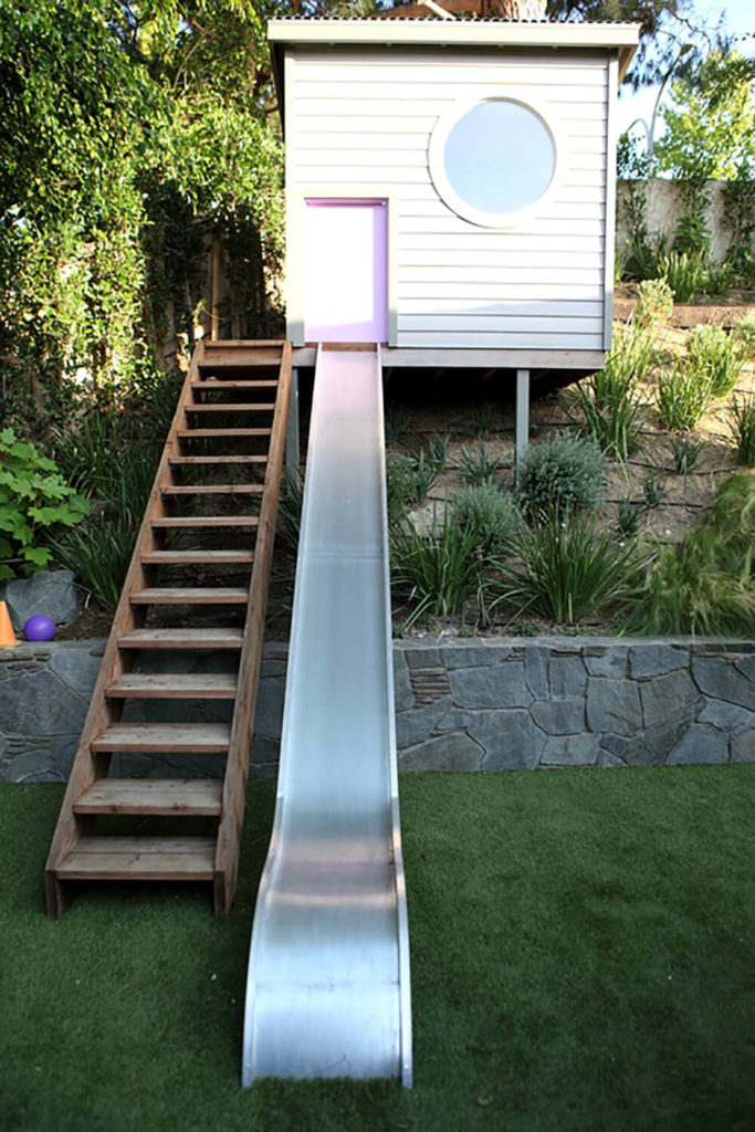 A long set of stairs leads up to a playhouse up on a hill. A metal slide leads down from a light pink door into the lush yard. A long set of stairs leads up to a playhouse up on a hill. A metal slide leads down from a light pink door into the lush yard.