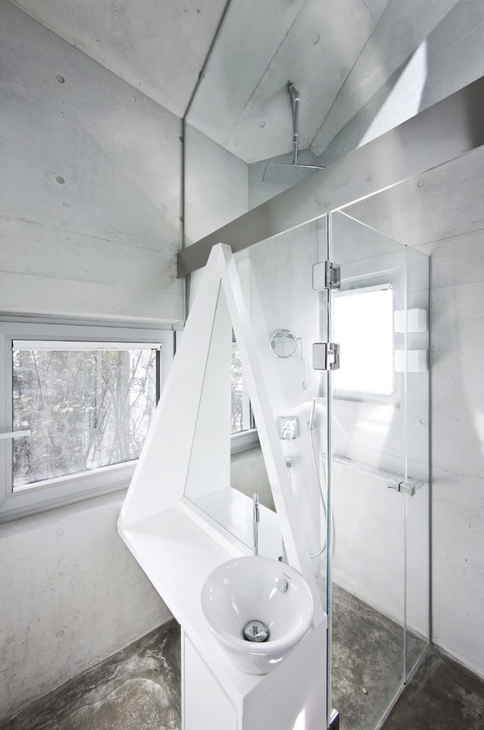 A white vessel sink fitted on a unique shaped vanity is fixed into the glass enclosure of the walk-in shower in this contemporary bathroom. It has concrete flooring and vaulted ceiling.