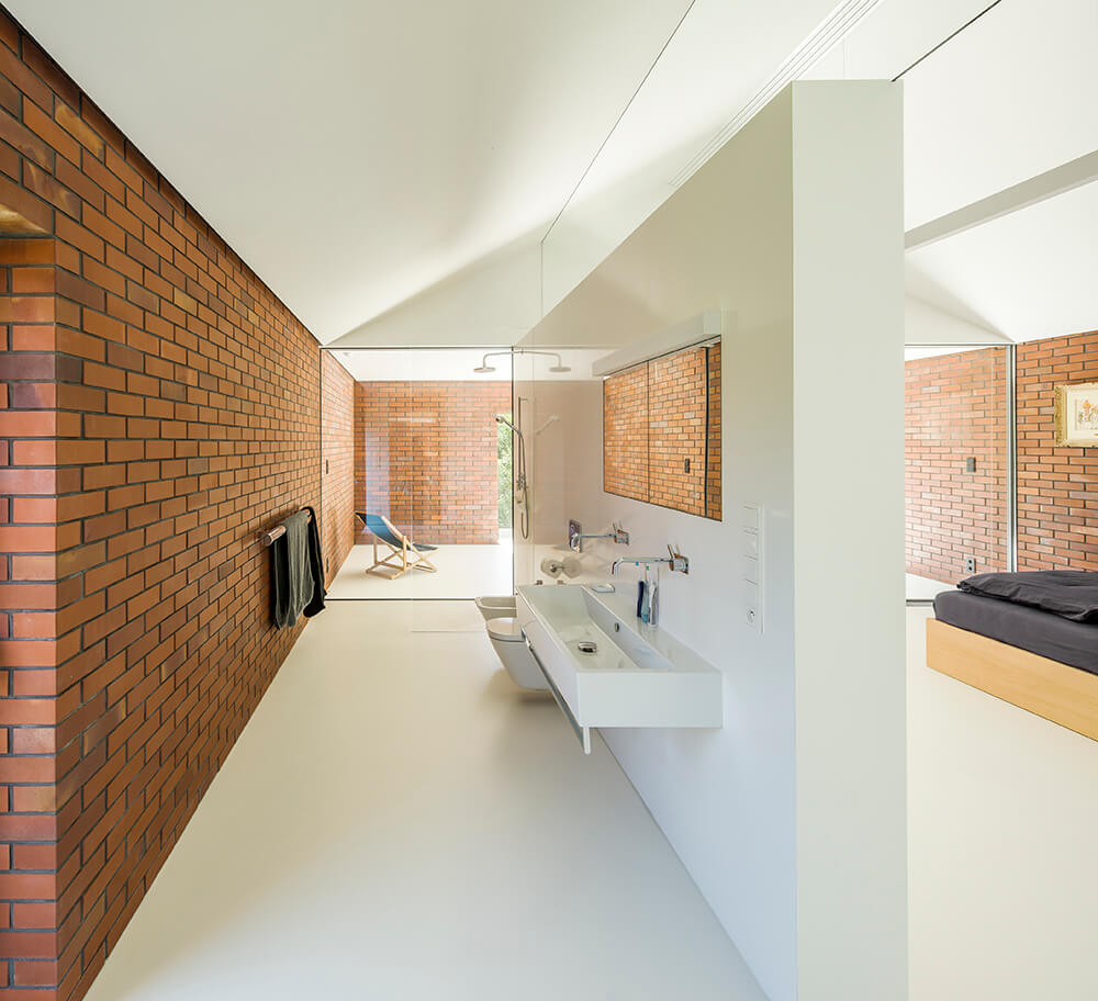 Galley bathroom with a left brick wall and white flooring. It includes an open shower area with a glass divider and wall mounted porcelain sink.