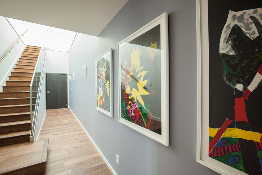Gray hallway designed with huge framed artworks and wood plank flooring that matches with the straight staircase framed with glass railing.