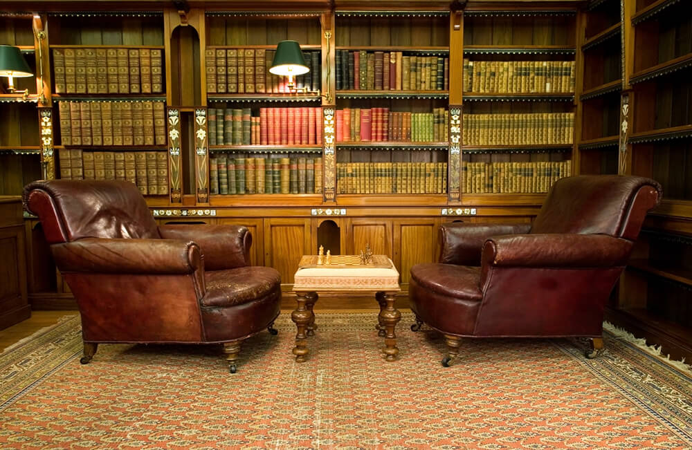 This home library with large bookshelves features a classy rug and brown leather seats with a chess board set as a centerpiece.
